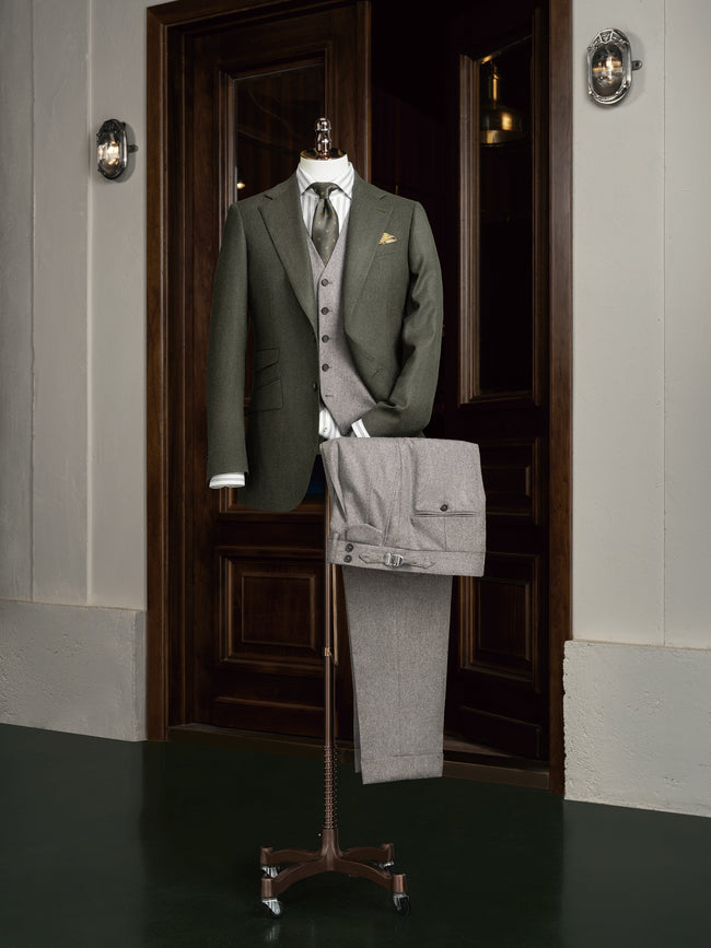 Complete formal outfit displayed on mannequin: olive green blazer, grey waistcoat, light grey flannel trousers, and striped shirt. A modern approach to timeless tailoring with tonal coordination.