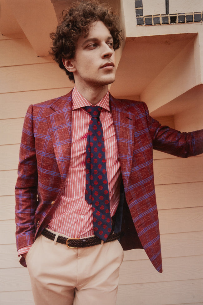 Full outfit view of a young man in a vibrant red and blue checked blazer, red striped shirt, navy tie with geometric pattern, and beige trousers. The look captures contemporary European tailoring with playful pattern layering.