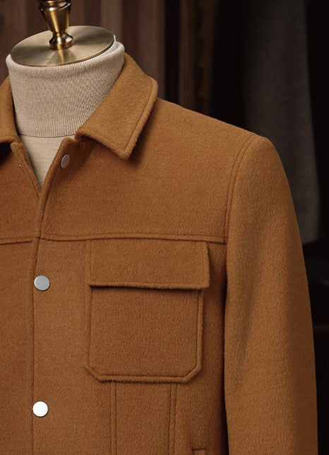Upper section of a camel wool aviator jacket with flap chest pockets, metal snap buttons, and a structured collar — layered over a cream roll-neck knit.
