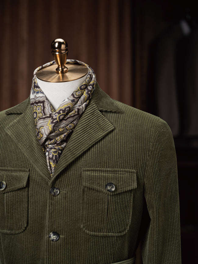 Upper body of a mannequin wearing an olive corduroy jacket with flap patch pockets, styled with a patterned paisley scarf — evoking vintage luxury with modern flair.