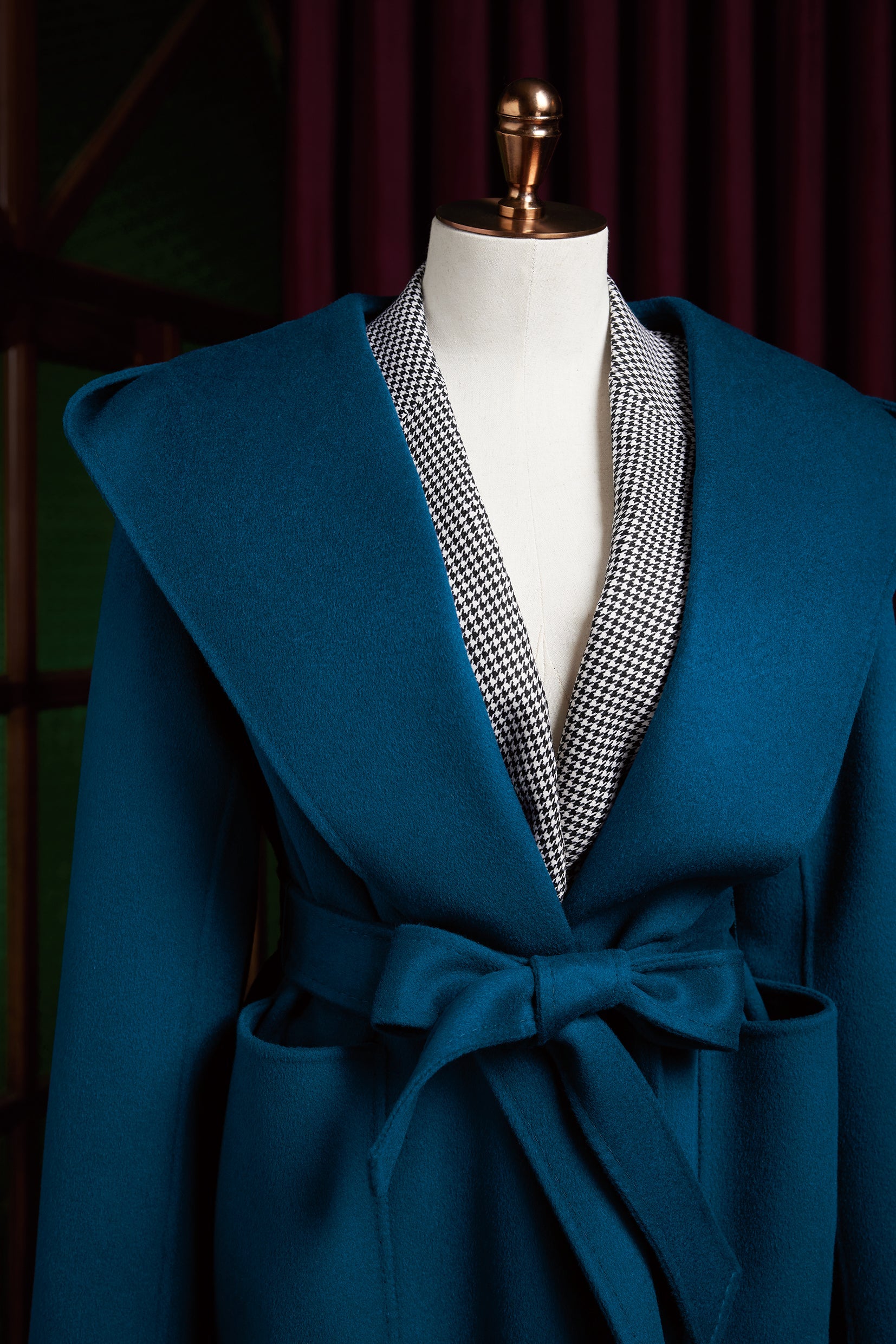 Close-up of a teal women's overcoat with a wide collar, belted waist, and houndstooth inner lining. The coat showcases a structured yet elegant design, perfect for formal or high-fashion wear