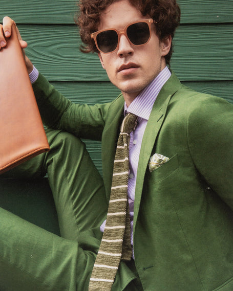 Man in a stylish green suit with a purple striped shirt and a green striped knit tie, holding a tan leather portfolio. He wears sunglasses and poses against a green textured background, exuding a high-fashion, contemporary look.