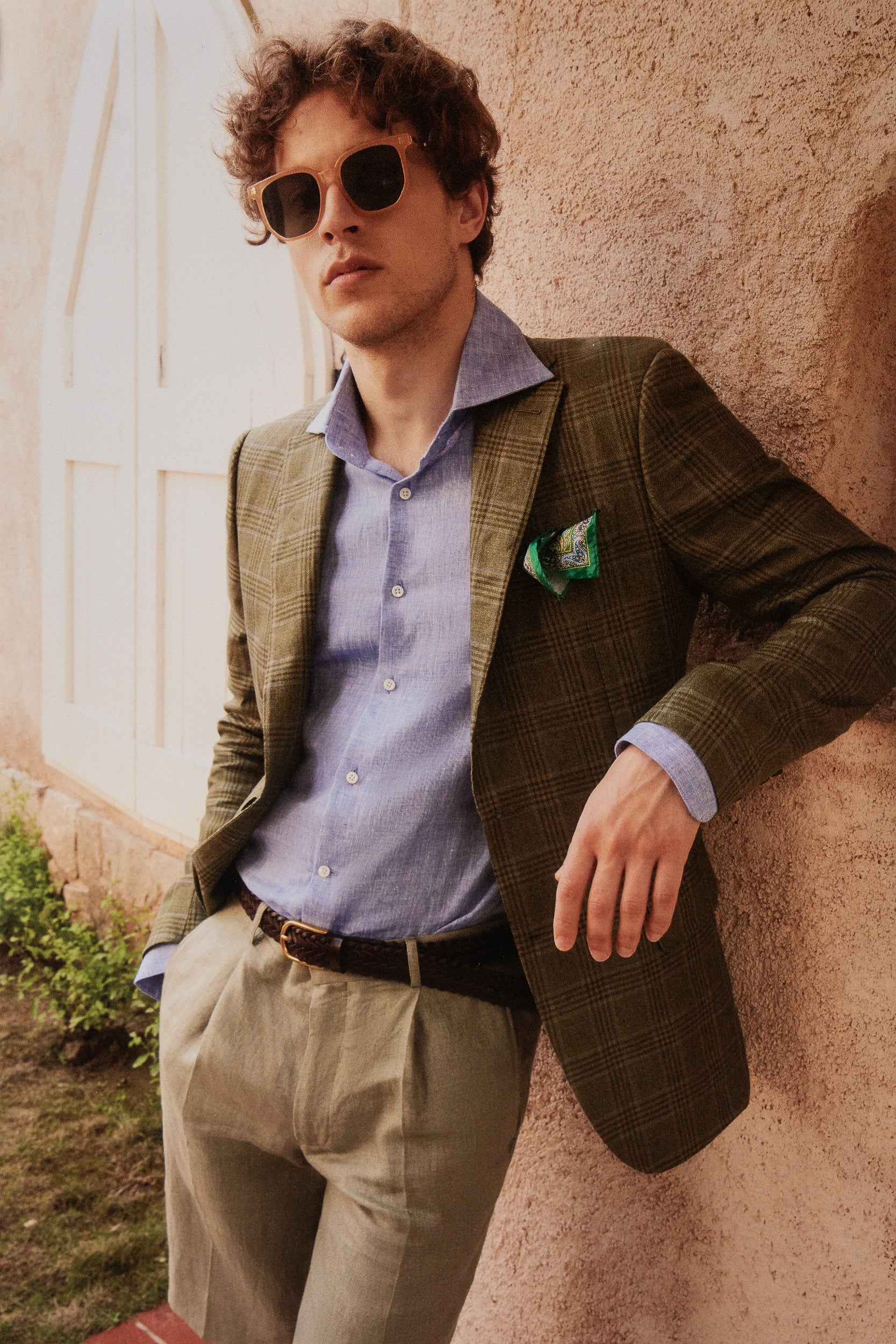 Model Wearing Brown Tailored Jacket with a Blue Undershirt and Beige trousers, Facing the camera leaning on a wall