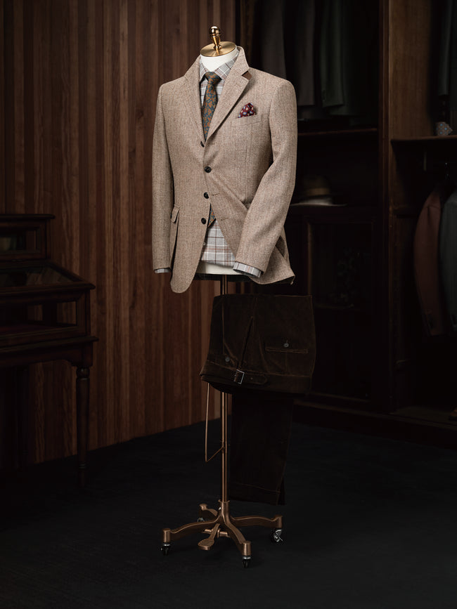 Elegant beige men's tweed blazer on a mannequin with a plaid shirt, floral tie, and pocket square, styled in a vintage-inspired tailoring shop.