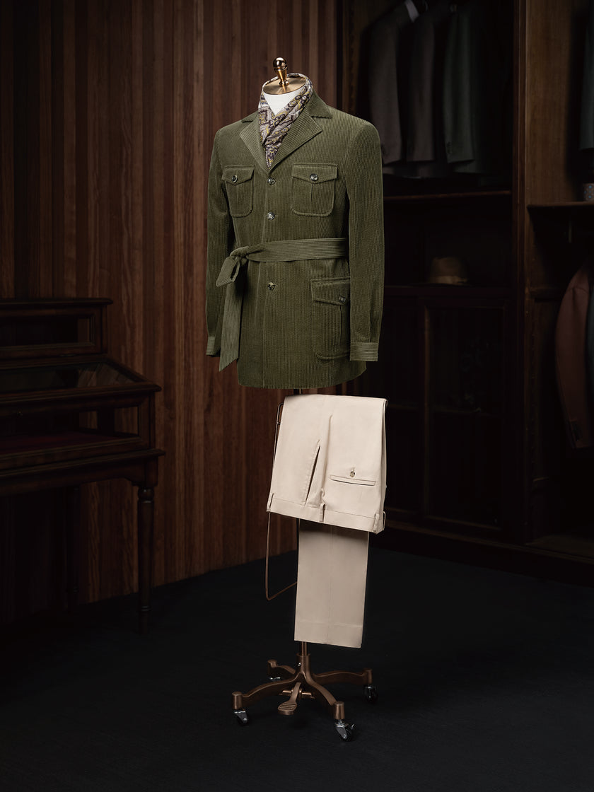 Full outfit presentation featuring an olive green corduroy belted jacket paired with beige cuffed trousers, styled on a mannequin in a vintage-inspired tailoring setting.