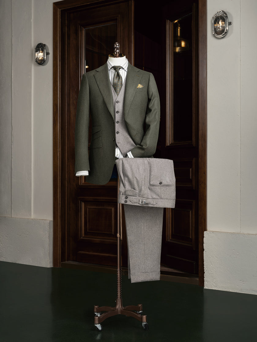 Complete formal outfit displayed on mannequin: olive green blazer, grey waistcoat, light grey flannel trousers, and striped shirt. A modern approach to timeless tailoring with tonal coordination.