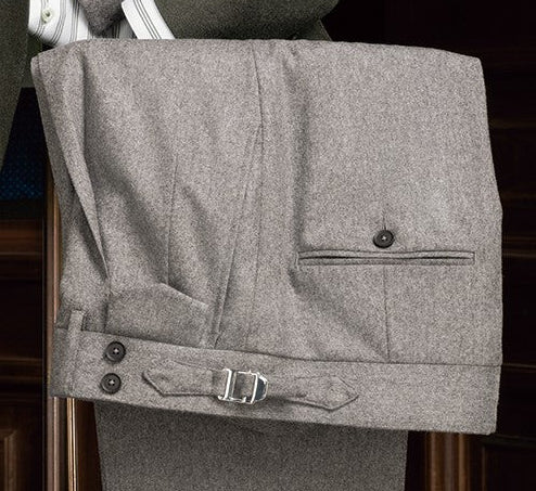 Folded light grey flannel trousers featuring side adjusters and a buttoned back pocket, highlighting traditional menswear tailoring with a modern fit.