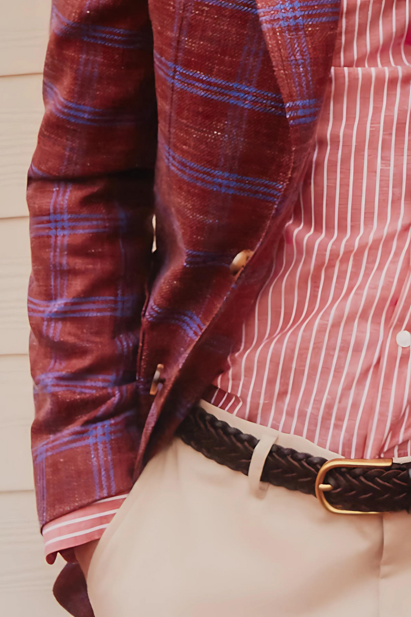 Close-up of a luxury red and blue checked linen blazer paired with a red striped dress shirt and beige trousers. The outfit features a braided dark leather belt and tortoiseshell buttons, highlighting refined tailoring and eclectic pattern mixing.