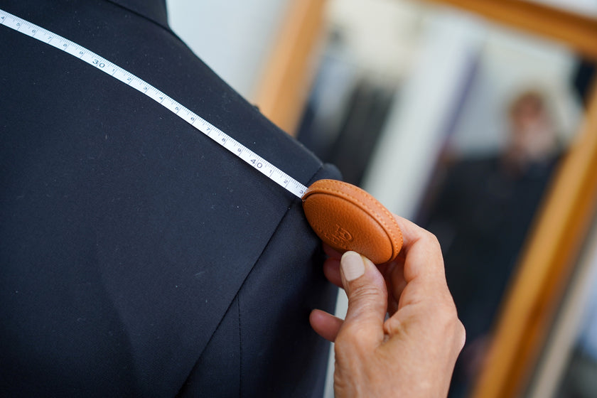 Close-up of a tailor taking a precise shoulder width measurement on a black tailored suit, using a luxury POLLHEIM measuring tape to ensure a perfect bespoke fit.