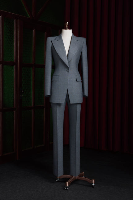 Full-length view of a tailored women’s grey power suit with structured shoulders and notch lapel, displayed on a mannequin against dark wood and curtain backdrop.
